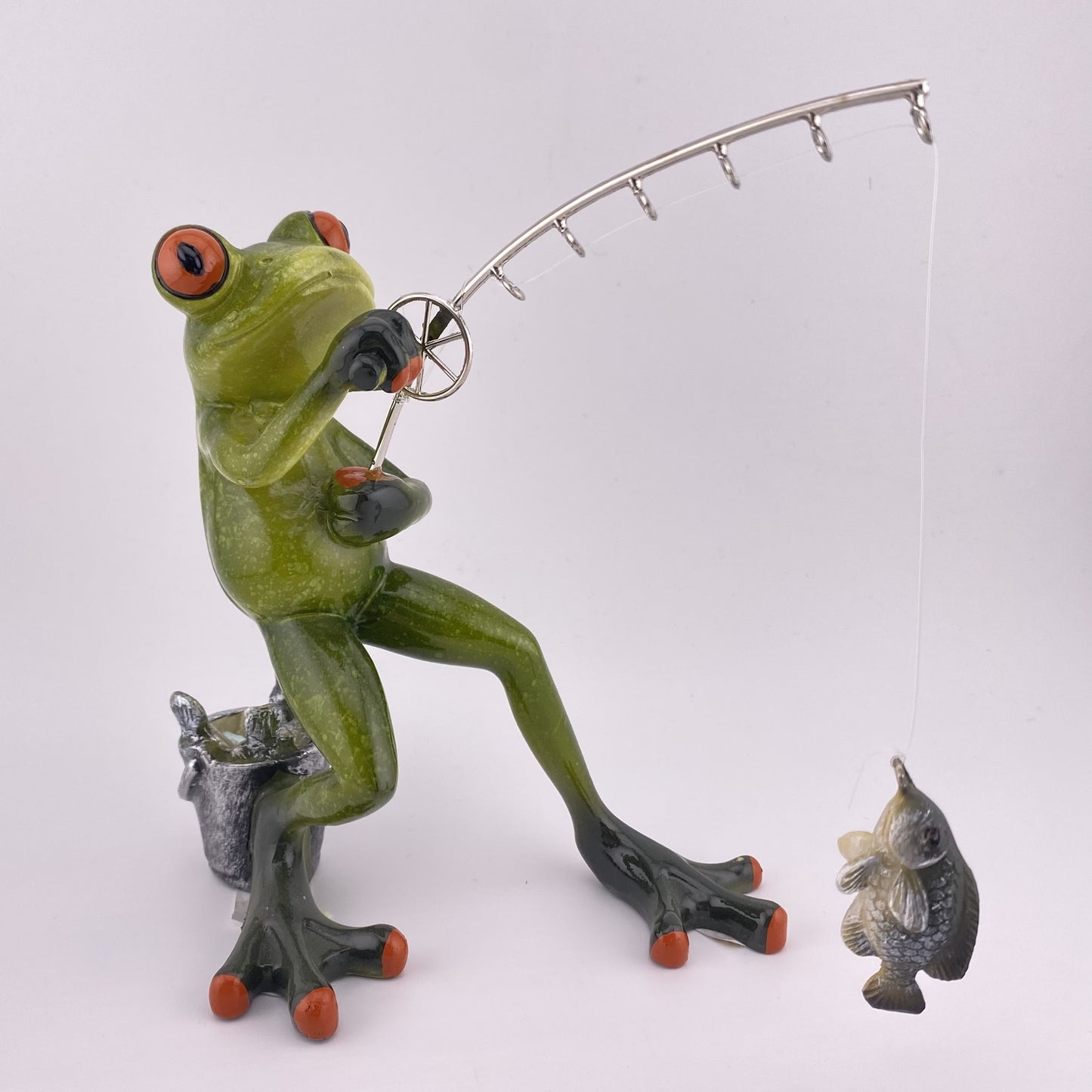 Eine Dekofigur vor weißem Hintergrund, die einen Frosch Mann beim Angeln abbildet. Der Deko Frosch steht und angelt mit all seiner Kraft einen Fisch, der am Haken seiner Angel hängt. 