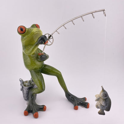 Eine Dekofigur vor weißem Hintergrund, die einen Frosch Mann beim Angeln abbildet. Der Deko Frosch steht und angelt mit all seiner Kraft einen Fisch, der am Haken seiner Angel hängt. 