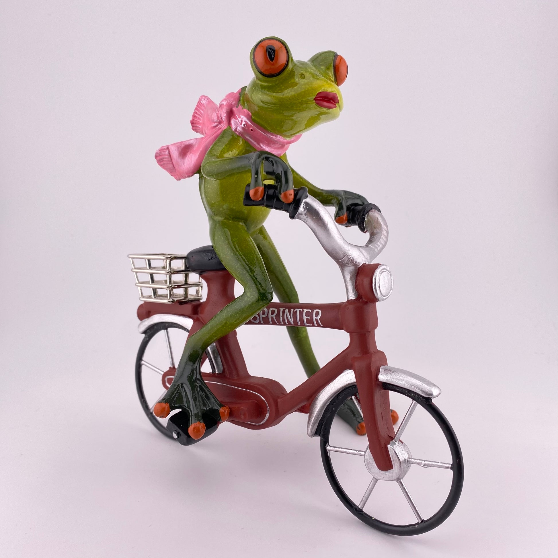 Eine Dekofigur vor weißem Hintergrund, die einen weiblichen Frosch auf einem rotem Fahrrad abbildet.