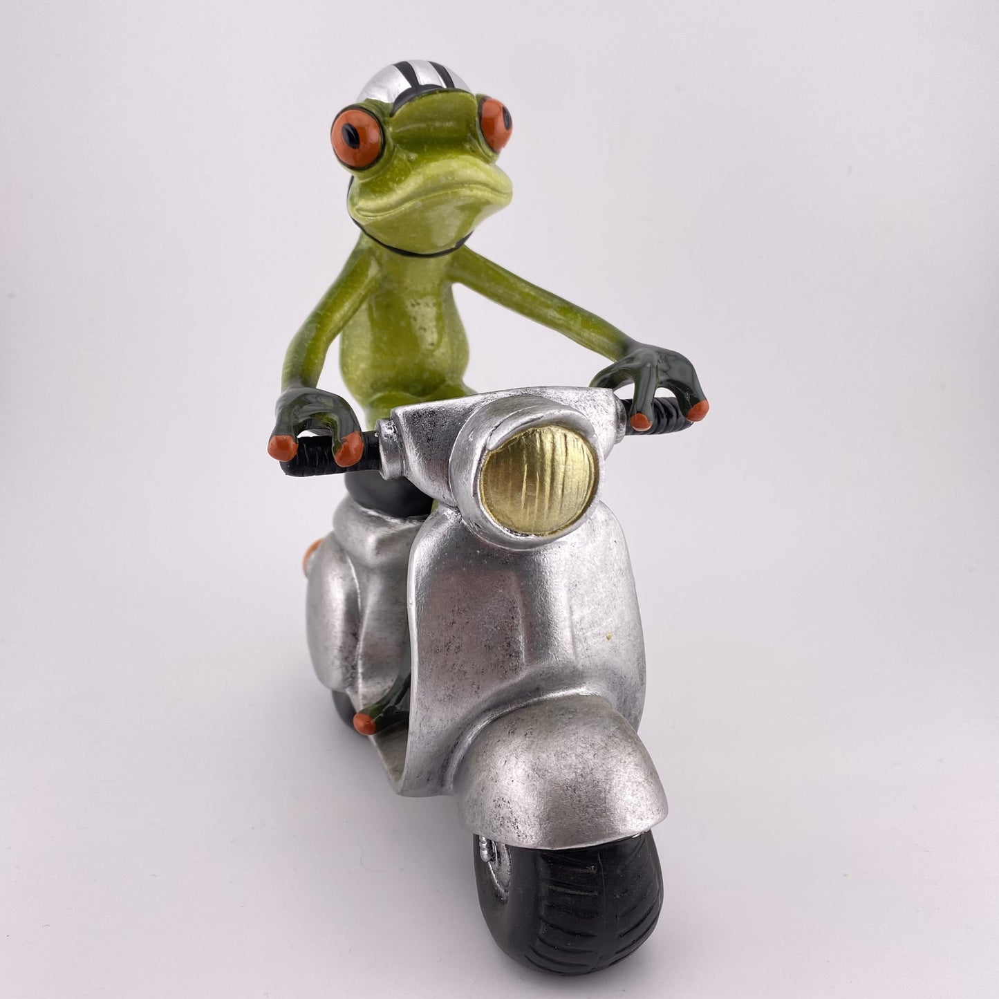 Eine Dekofigur vor weißem Hintergrund, die einen männlichen Frosch auf einem silbernen Roller abbildet.