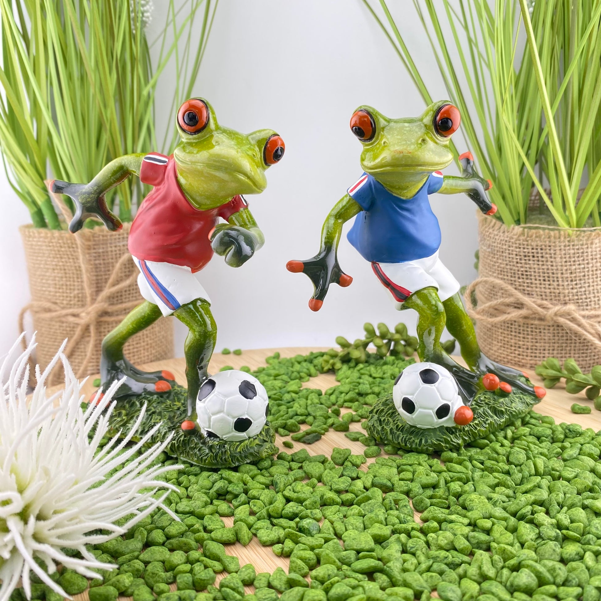 Zwei Frosch Dekofiguren im Ambiente, die fußballspielende Frösche in rotem und blauem Trikot mit Fußball abbildet.