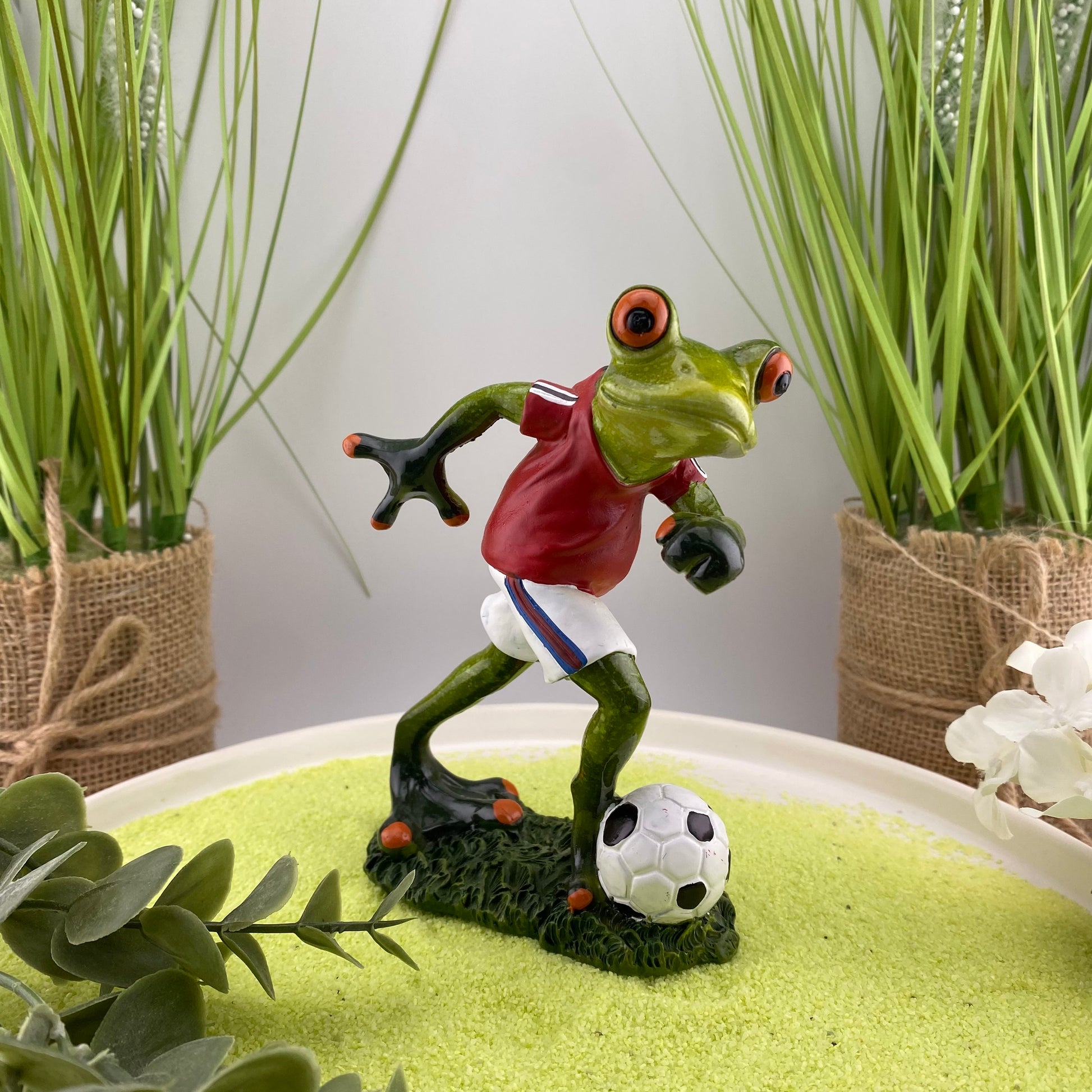 Eine Dekofigur im Ambiente, die einen fußballspielenden Frosch in rotem Trikot mit Fußball abbildet.