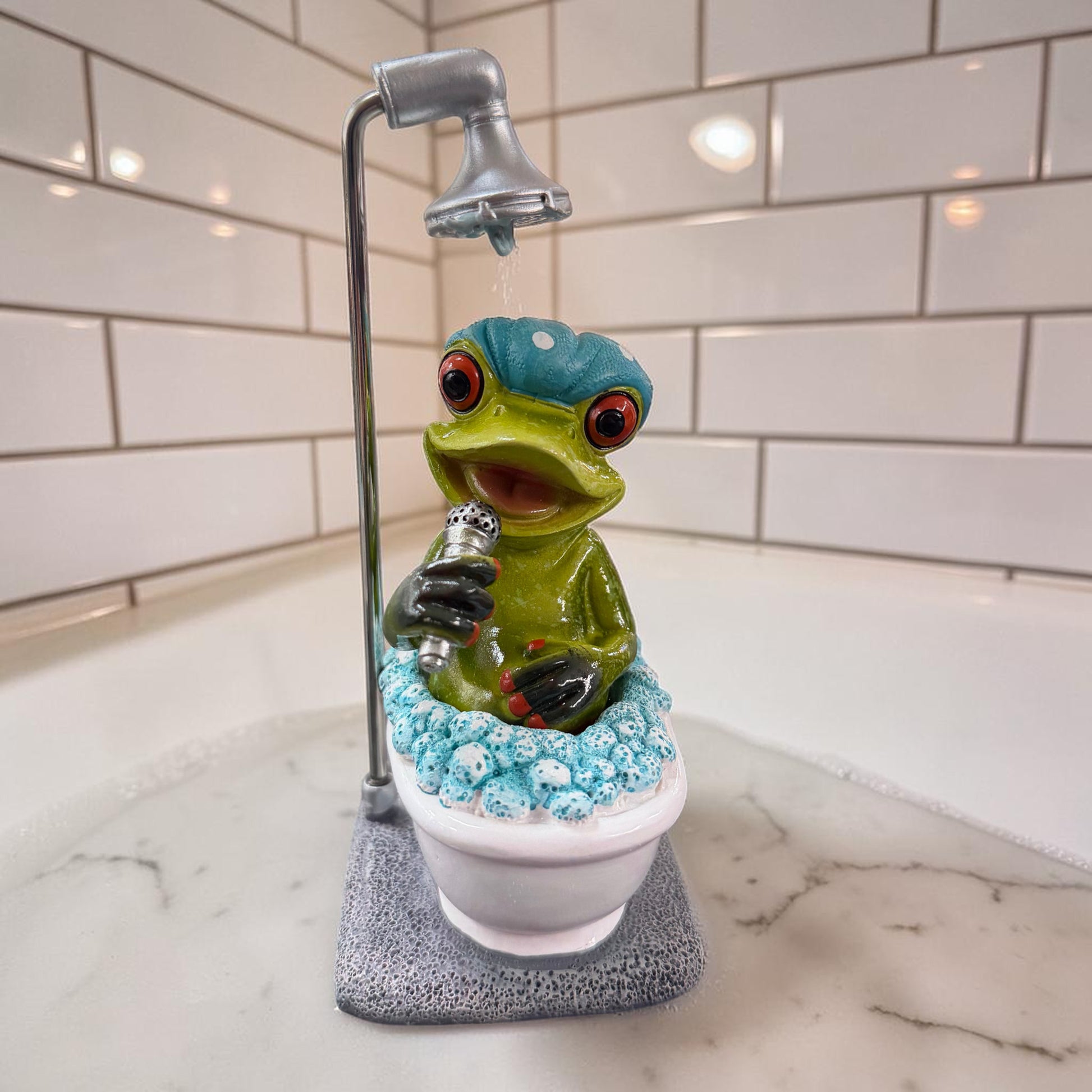 Grüner Formano Frosch mit blauer Badehaube und Mikrofon sitzt unter einer Dusche in einer weißen Badewanne vor weißen Fliesen.