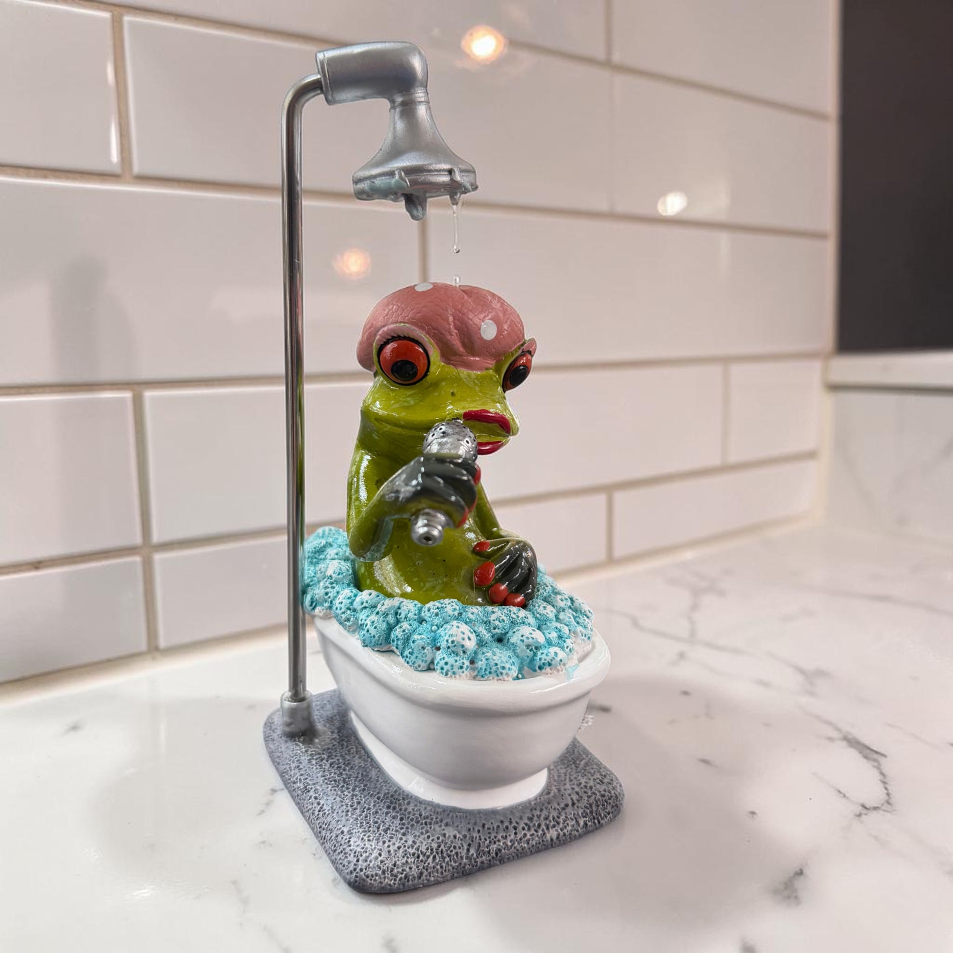 Grüner Formano Frosch mit rosa Badehaube und Mikrofon sitzt unter einer Dusche in einer weißen Badewanne vor weißen Fliesen.