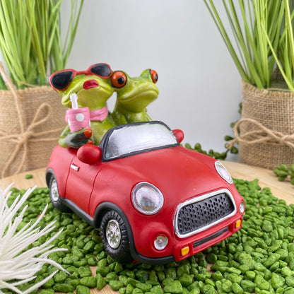 Ein Dekofigur Froschpaar der Serie Froschhausen sitzt in einem rotem Auto Cabrio. Die Frosch-Dame trägt eine Sonnenbrille und trinkt ein Kaltgetränk. Der Frosch-Mann fährst das Auto. Die Frosch-Dekofigur ist im Ambiente platziert.