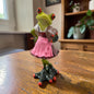 Grüner Formano Frosch im rosa Dirndl mit einem braunen Mausi-Herz auf einem Holztisch.