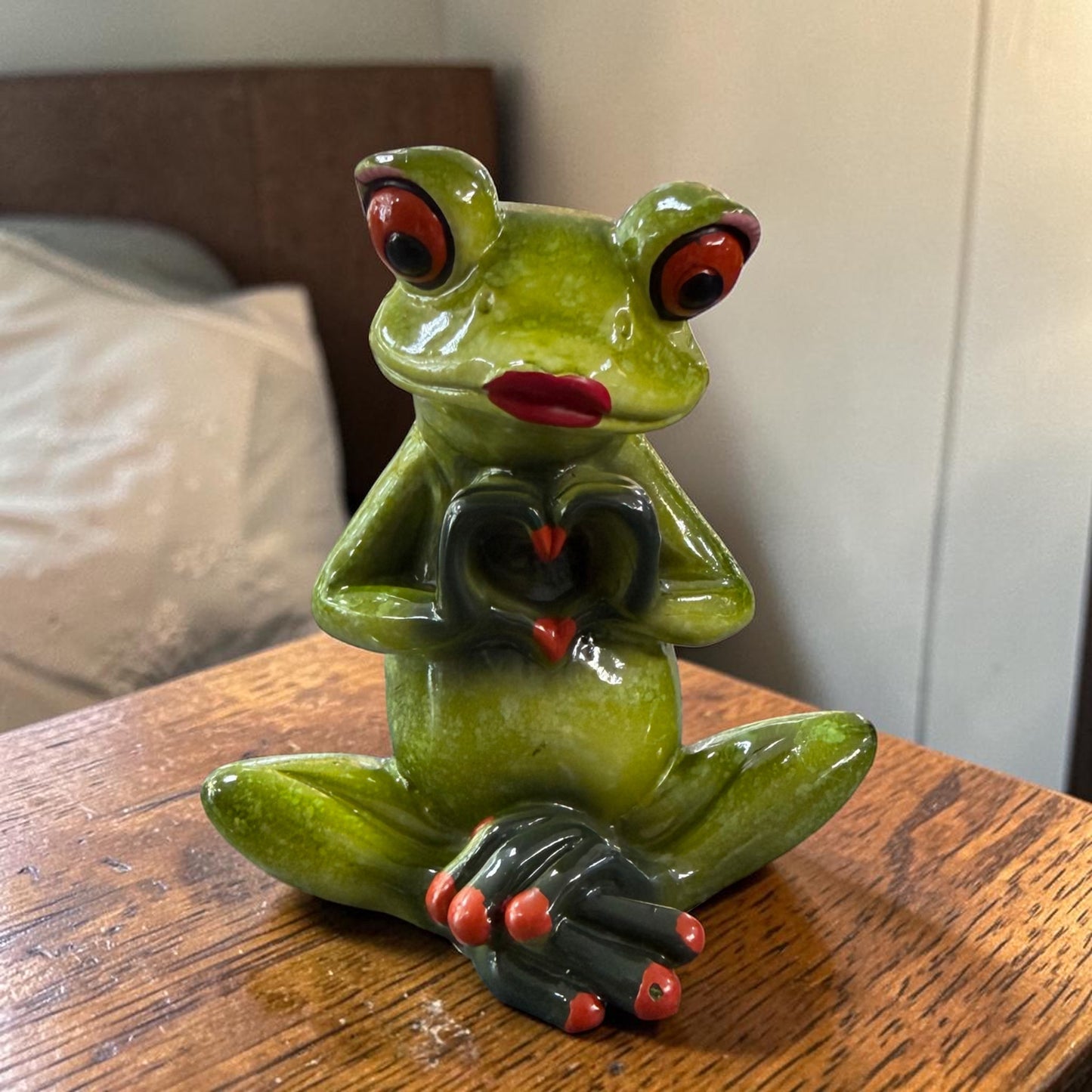 Sitzender grüner Formano Frosch mit roten Lippen formt mit seinen dunklen Händen ein Herz auf einem Holztisch.