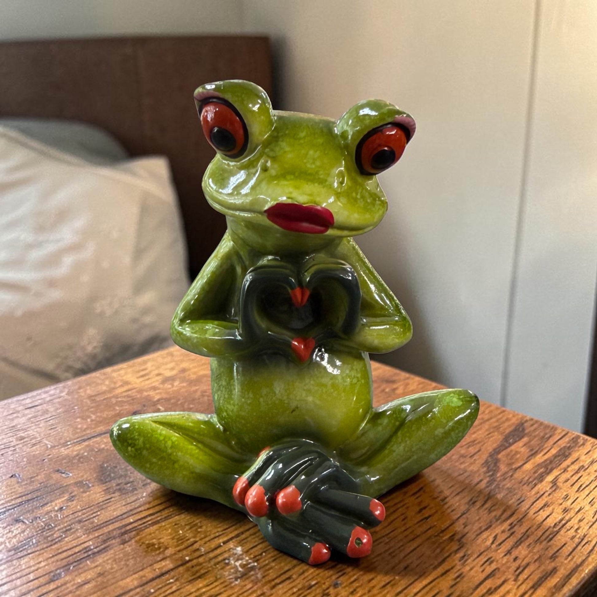 Sitzender grüner Formano Frosch mit roten Lippen formt mit seinen dunklen Händen ein Herz auf einem Holztisch.