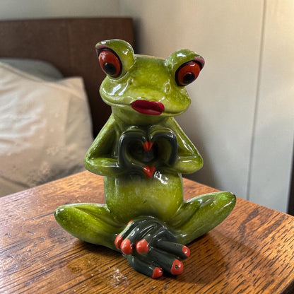 Sitzender grüner Formano Frosch mit roten Lippen formt mit seinen dunklen Händen ein Herz auf einem Holztisch.