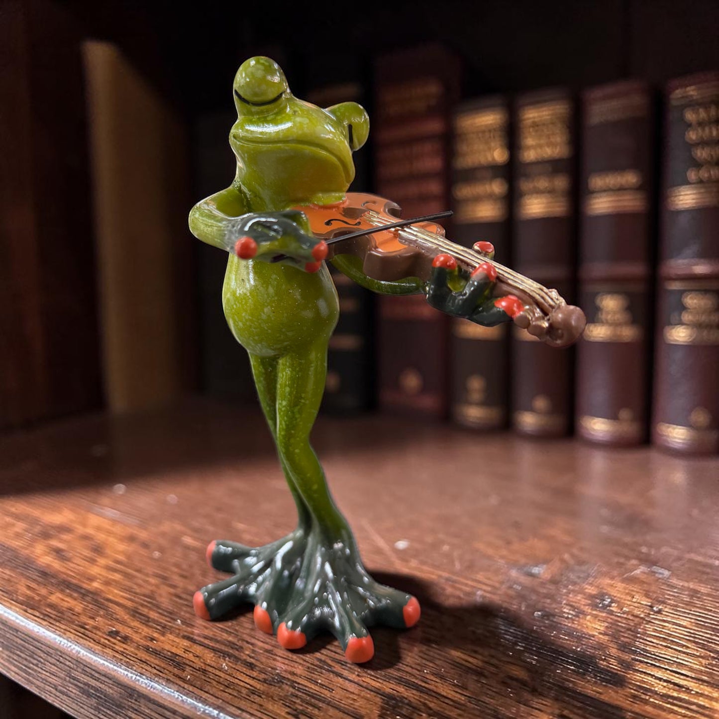 Grüner Formano Frosch mit Geige auf einem dunklen Holzregal vor Büchern.