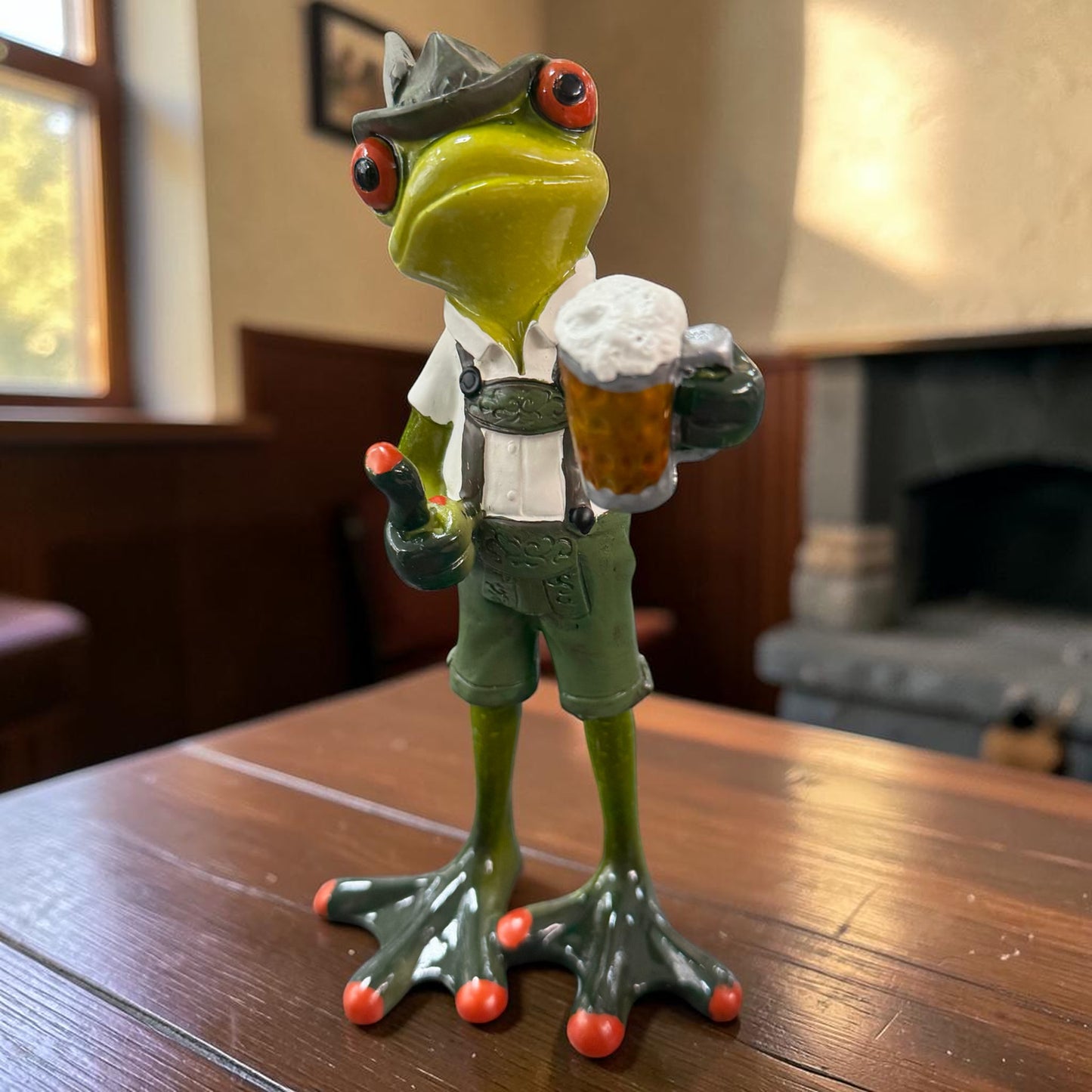Grüner Formano Frosch in Lederhose mit Bierkrug auf einem Holztisch in einem Innenraum.