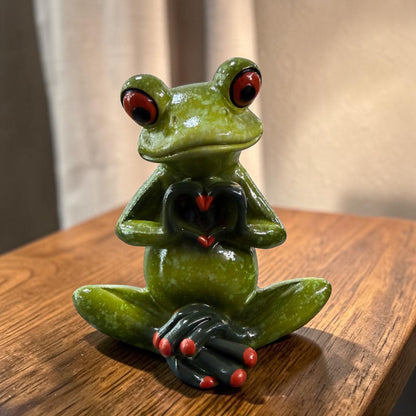 Sitzender grüner Formano Frosch formt mit seinen dunklen Händen ein Herz auf einem hellbraunen Holztisch.