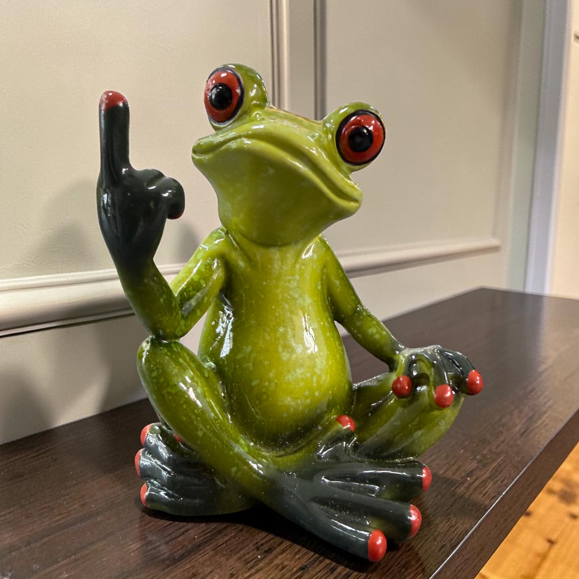 Sitzender grüner Formano Frosch zeigt einen erhobenen Mittelfinger auf einem dunklen Holzregal vor einer hellen Wand.