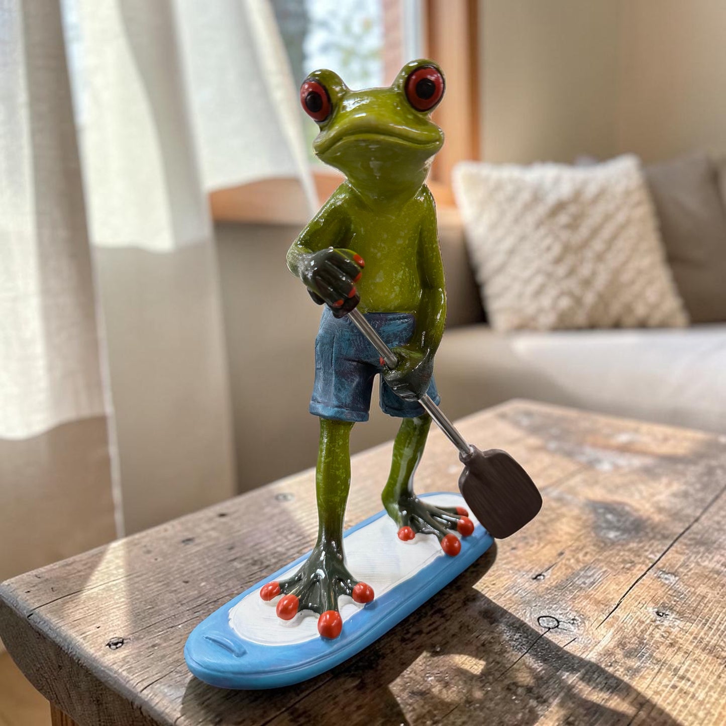 Grüner Formano Frosch in blauer Badehose steht mit einem Paddel auf einem blau-weißen Paddleboard auf einem rustikalen Holztisch.