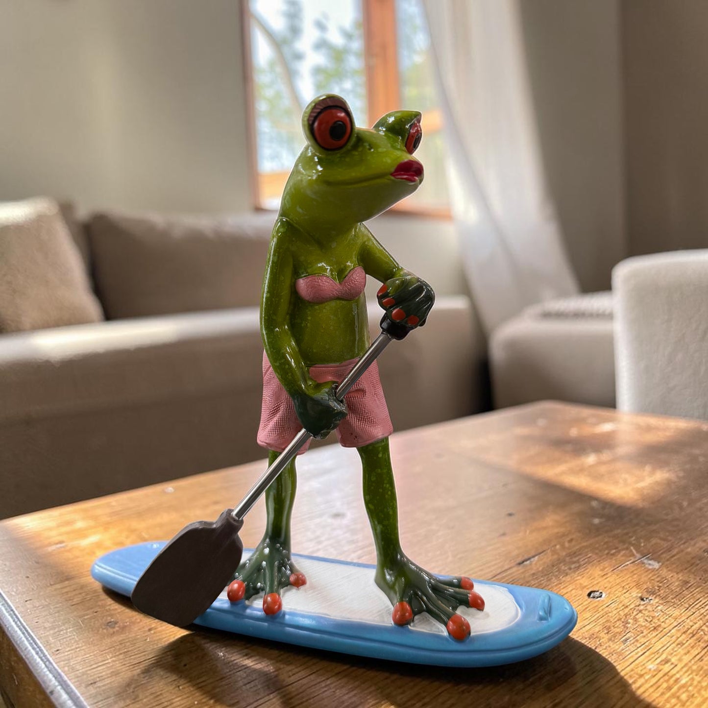 Grüner Formano Frosch im rosa Bikini steht mit einem Paddel auf einem blau-weißen Paddleboard auf einem Holztisch.