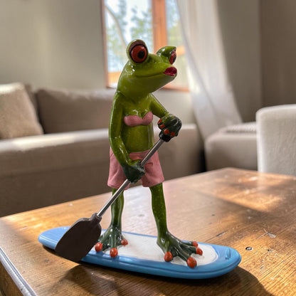 Grüner Formano Frosch im rosa Bikini steht mit einem Paddel auf einem blau-weißen Paddleboard auf einem Holztisch.
