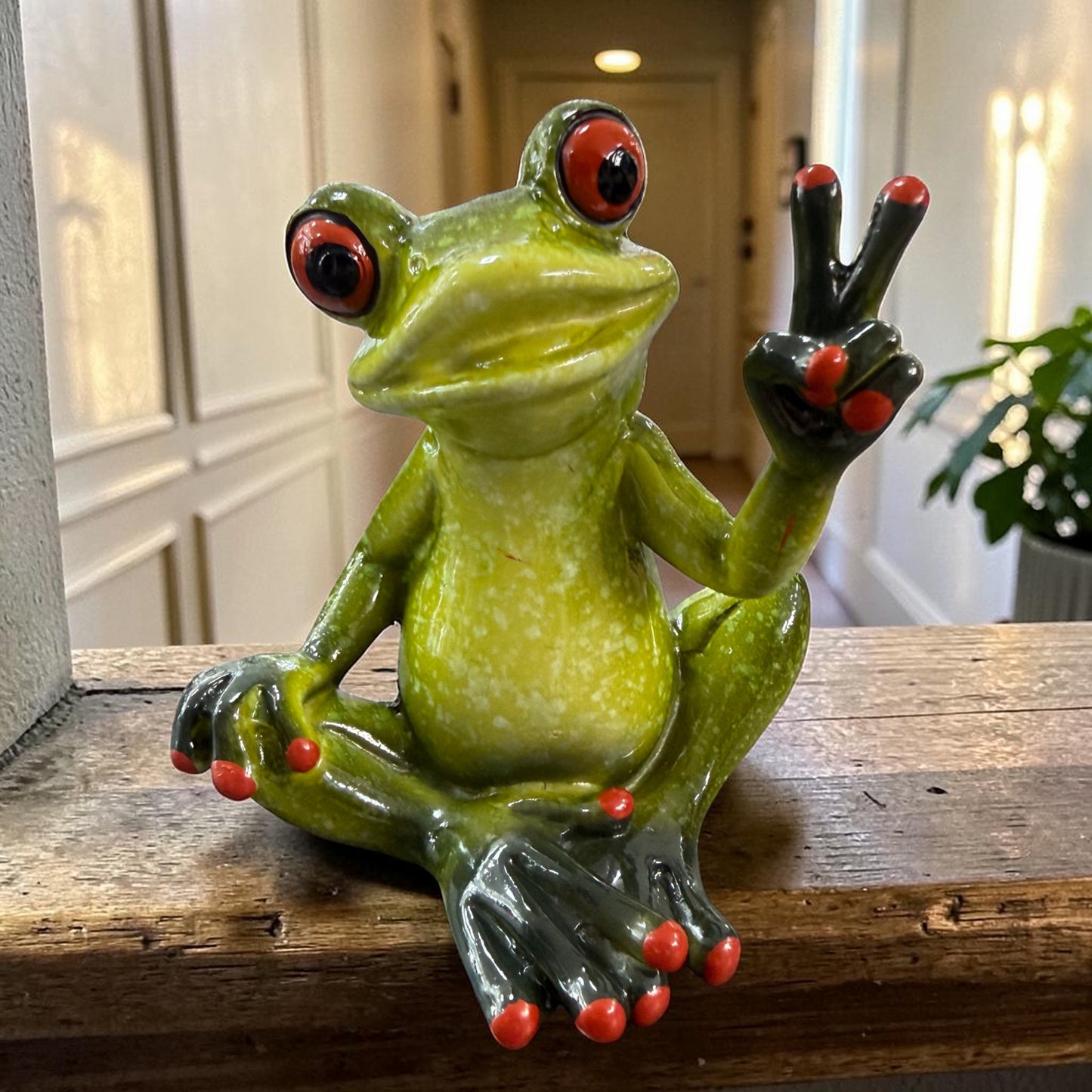 Sitzender grüner Formano Frosch zeigt mit einer Hand ein Peace-Zeichen auf einem rustikalen Holzregal.