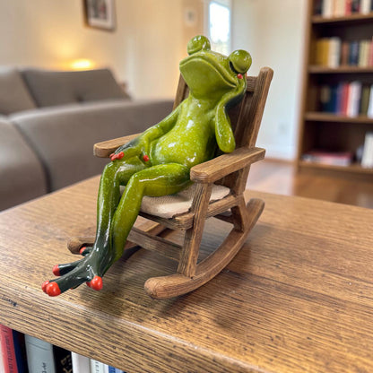 Grüner Formano Frosch sitzt mit geschlossenen Augen in einem braunen Schaukelstuhl auf einem Holztisch.