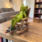 Grüner Formano Frosch sitzt mit geschlossenen Augen in einem braunen Schaukelstuhl auf einem Holztisch.