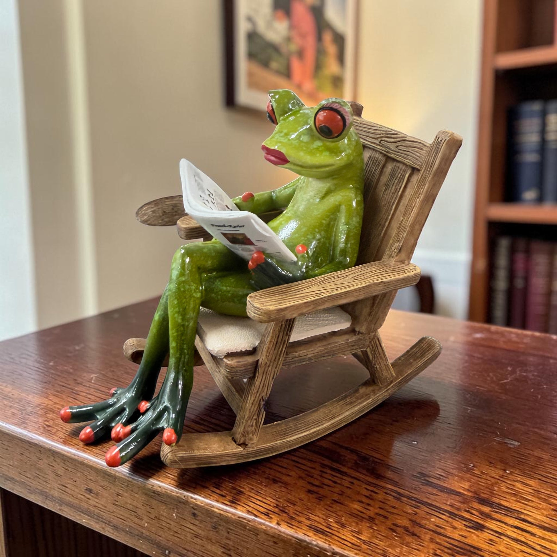 Grüner Formano Frosch sitzt mit einer Zeitung in einem braunen Schaukelstuhl auf einem dunklen Holztisch.