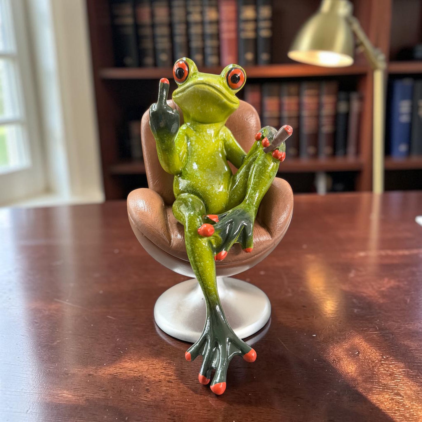 Grüner Formano Frosch sitzt mit Zigarre und erhobenem Mittelfinger in einem braunen Drehsessel auf einem Holztisch.
