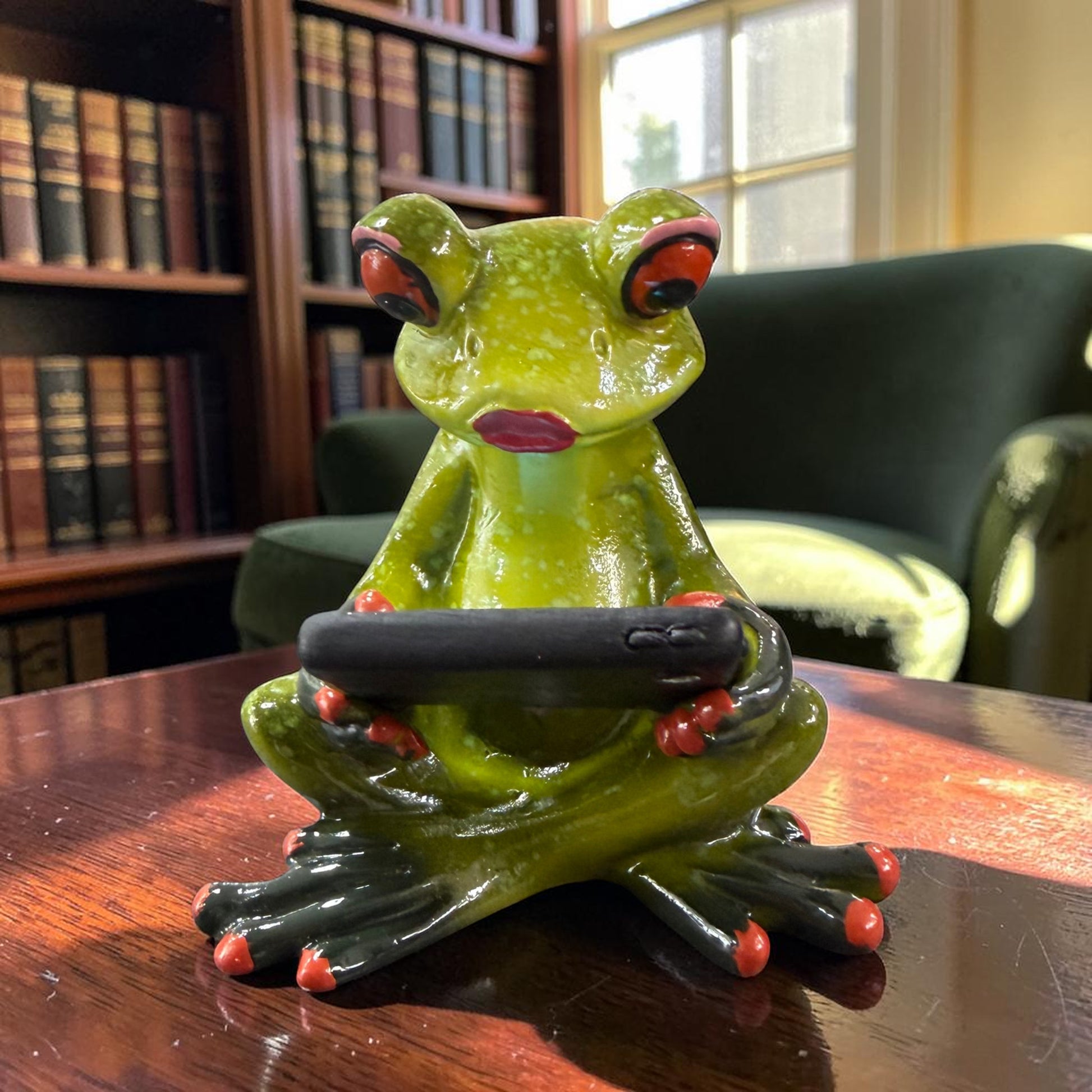 Grüner Formano Frosch sitzt mit gekreuzten Beinen und hält ein schwarzes Tablet auf einem dunklen Holztisch vor einem Bücherregal.
