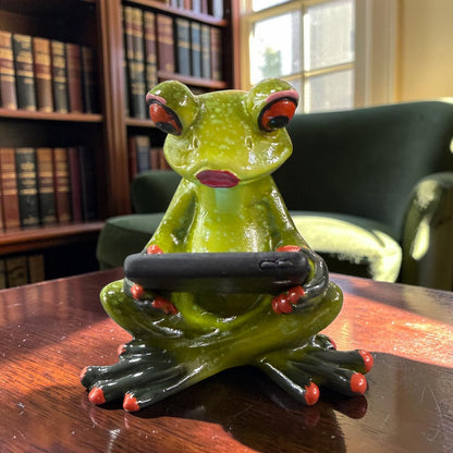 Grüner Formano Frosch sitzt mit gekreuzten Beinen und hält ein schwarzes Tablet auf einem dunklen Holztisch vor einem Bücherregal.