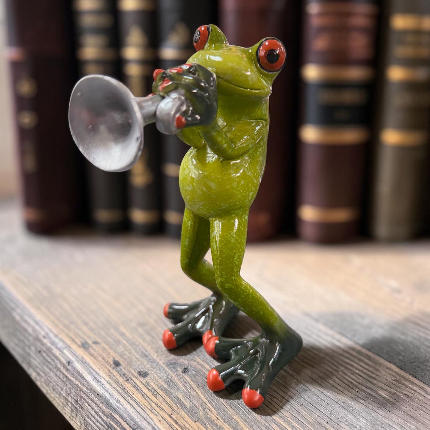 Grüner Formano Frosch mit silberner Trompete auf einem Holzregal vor Büchern.