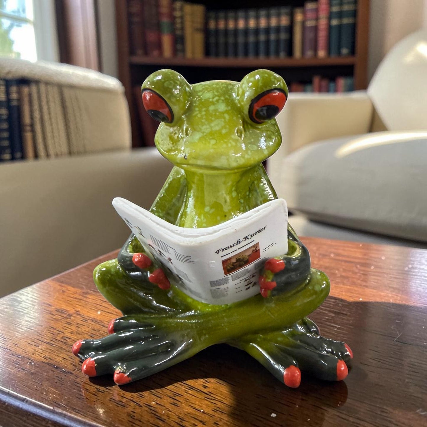 Grüner Formano Frosch sitzt mit gekreuzten Beinen und liest den weißen Frosch-Kurier auf einem dunklen Holztisch.