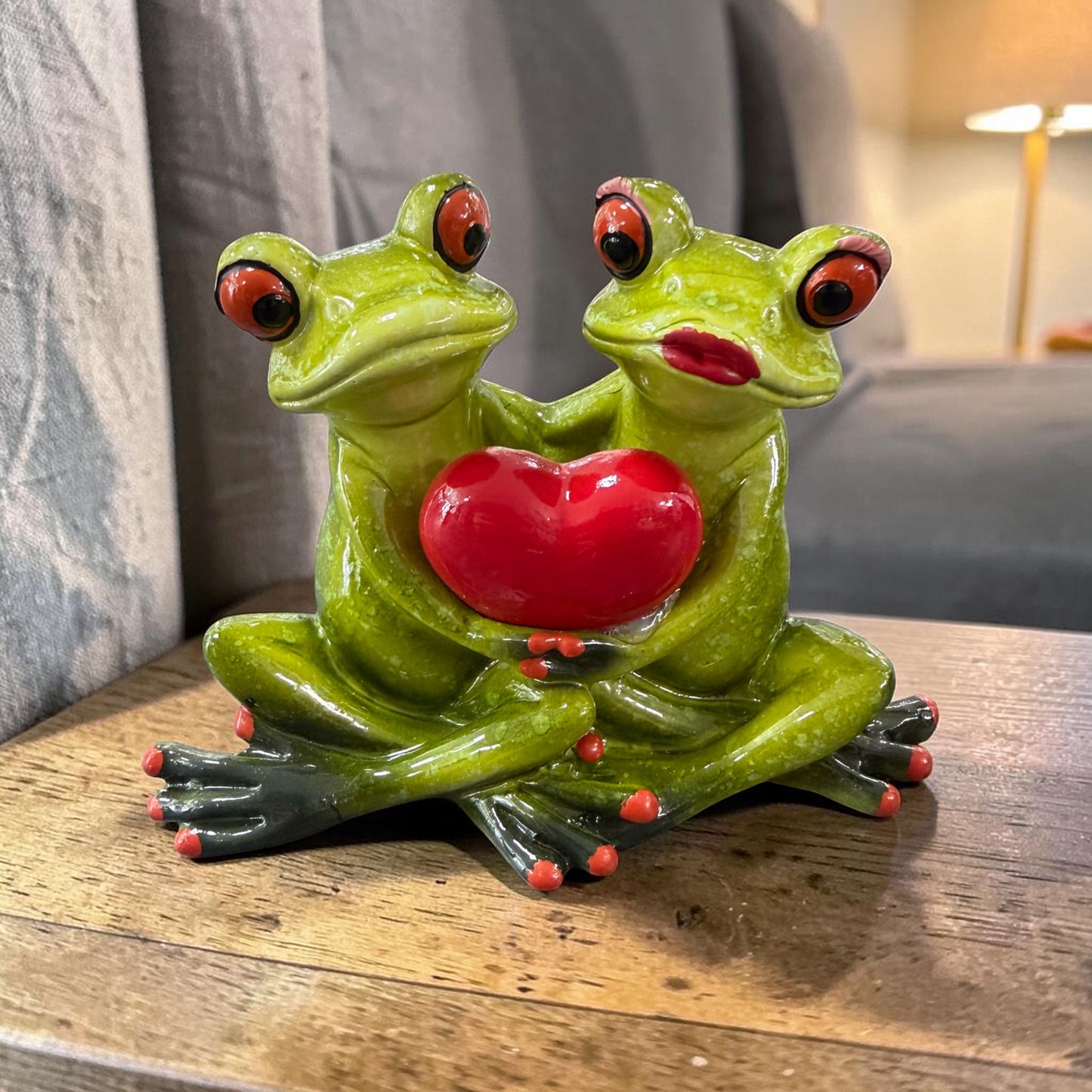 Zwei sitzende grüne Formano Frösche halten gemeinsam ein rotes Herz auf einem Holztisch vor einem grauen Sofa.
