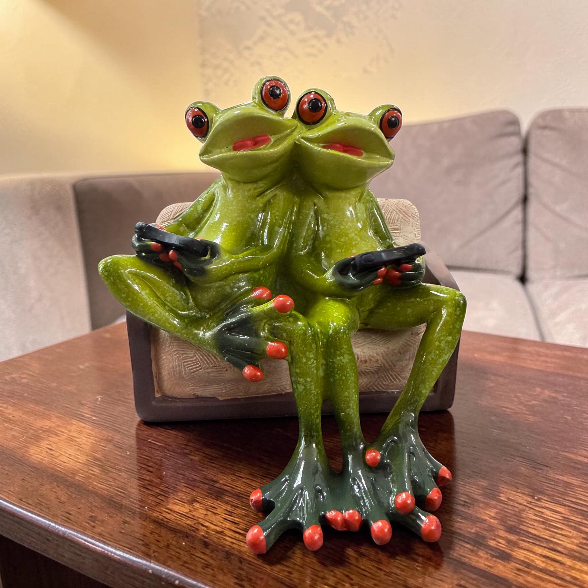 Zwei grüne Formano Frösche sitzen mit schwarzen Fernbedienungen auf einem braunen Sofa auf einem Holztisch.