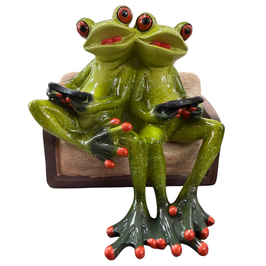 Zwei grüne Formano Frösche sitzen mit schwarzen Fernbedienungen auf einem braunen Sofa vor weißem Hintergrund.