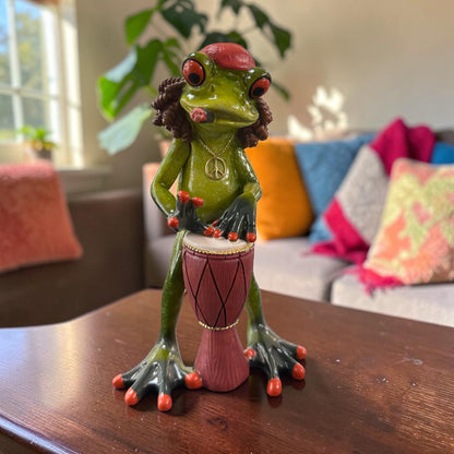Grüner Formano Hippie-Frosch mit roter Mütze, Peace-Kette und Trommel steht auf einem dunklen Holztisch.