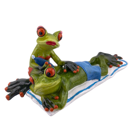 Ein Dekofigur Froschpaar der Serie Froschhausen, bei dem die Frosch Frau den Frosch Mann eincremt, während er auf einem Handtuch liegt. Die Sommerdekoration ist vor weißem Hintergrund platziert.