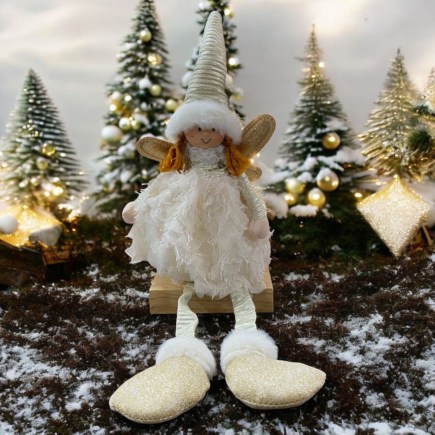 Eine sitzende Stoff-Engelfigur mit langer Zipfelmütze, goldenen Flügeln und flauschigem Kleid in der Farbe Beige im Ambiente