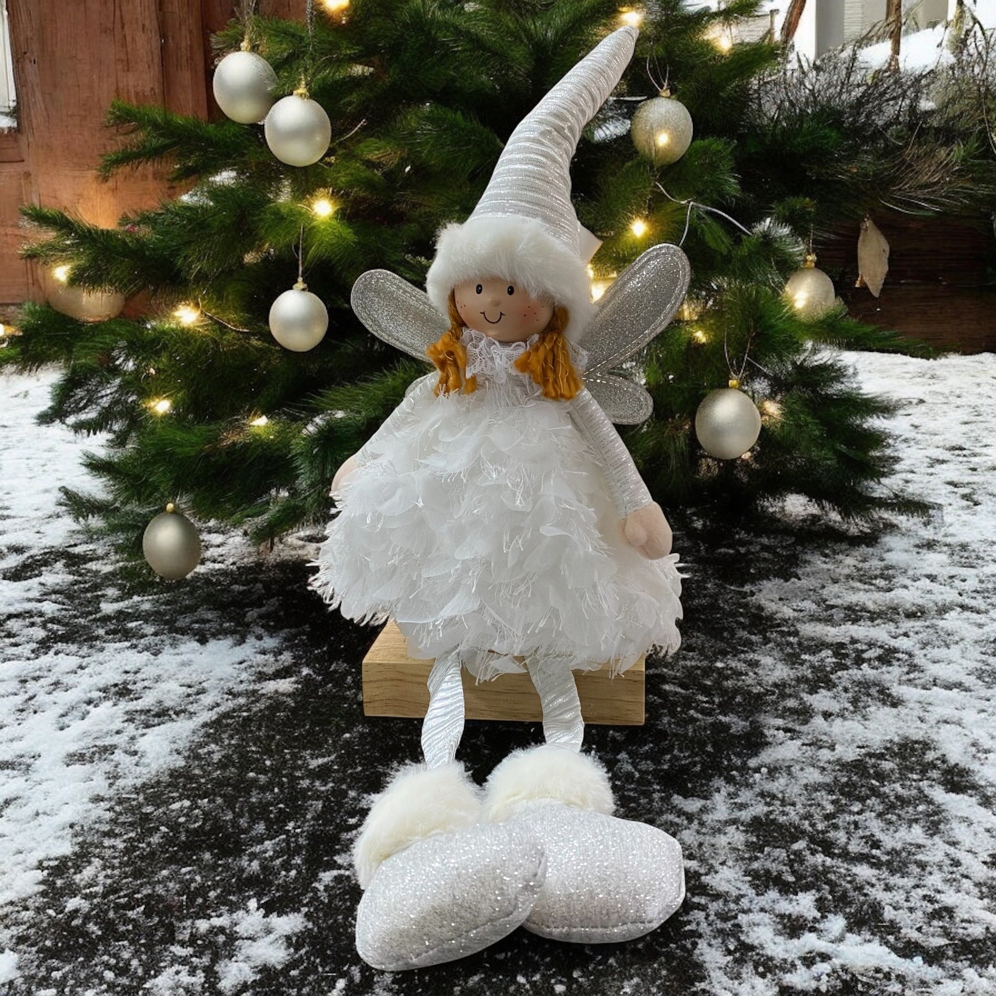 Eine sitzende Stoff-Engelfigur mit langer Zipfelmütze, silbernen Flügeln und flauschigem Kleid in der Farb Weiß im Ambiente