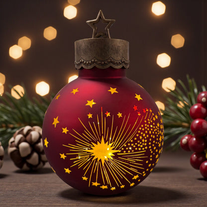 Eine dunkelrote Dekokugel mit goldfarbenem Stern- und Strahlenmuster steht beleuchtet auf einem Tisch, umgeben von Tannenzweigen, Beeren und unscharfen Lichtern im Hintergrund.