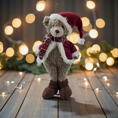 Dekofigur in Form eines Teddys mit beigem Fell, bekleidet mit roter Mütze mit weißem Rand, rotem Schal mit schwarzen und weißen Mustern sowie roter Jacke mit weißem Besatz, stehend auf brauner Holzoberfläche vor unscharfem Hintergrund mit warmgelben Lichtern und grünen Elementen.