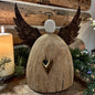 Eine größere Dekofigur in Form eines Engels aus hellbraunem Holz mit ausgeprägter Struktur, dunklen Metallflügeln und weißem Kopf, auf Holzoberfläche vor braunem Holzhintergrund, daneben eine weiße Kerze mit gelblicher Flamme und grüne Zweige.