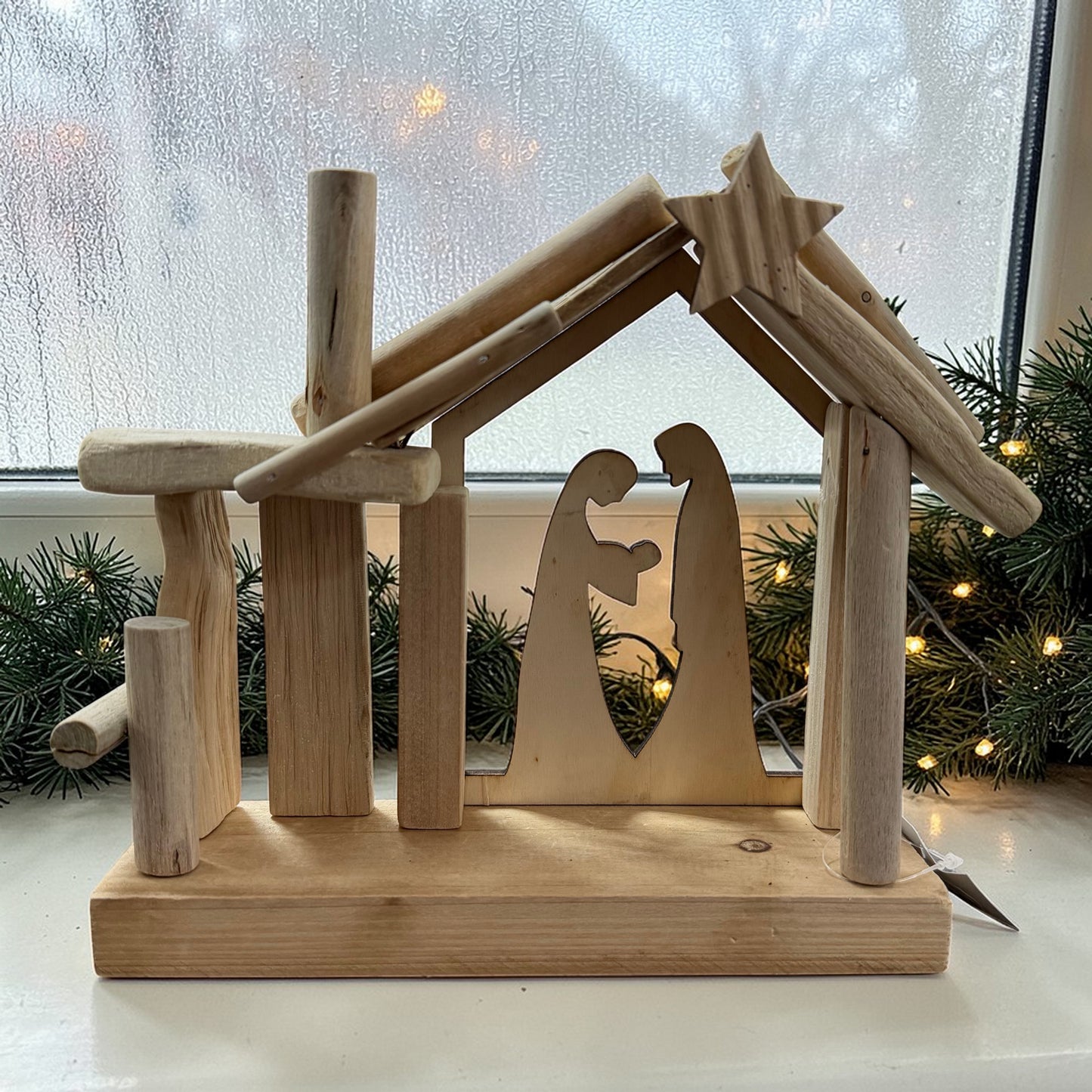 Dekorative Holzkrippe mit ausgeschnittener Darstellung der Heiligen Familie auf einer Fensterbank. Im Hintergrund sorgen Tannenzweige und warme Lichter für eine ruhige, weihnachtliche Stimmung.