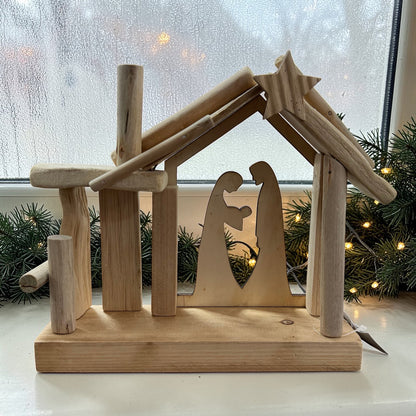 Dekorative Holzkrippe mit ausgeschnittener Darstellung der Heiligen Familie auf einer Fensterbank. Im Hintergrund sorgen Tannenzweige und warme Lichter für eine ruhige, weihnachtliche Stimmung.