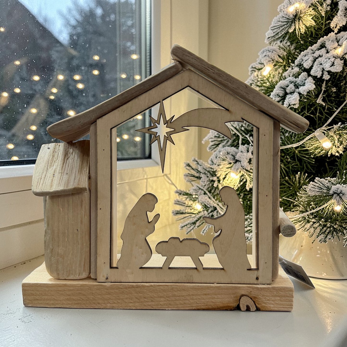 Dekorative Holzkrippe mit Sternschnuppen-Motiv auf einer Fensterbank arrangiert. Im Hintergrund sorgen Lichter und winterliche Tannenzweige für eine stimmungsvolle Weihnachtsatmosphäre.