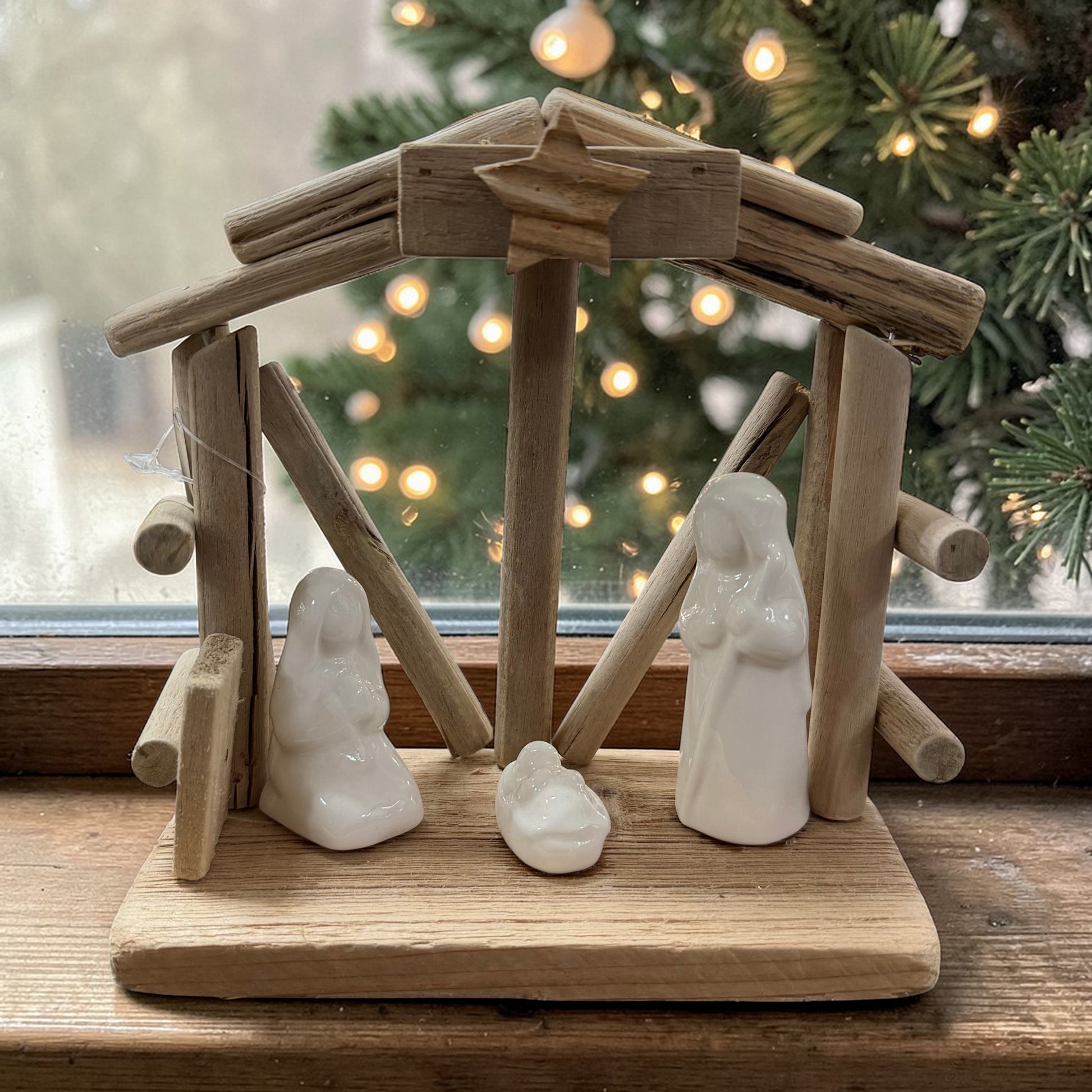 Holzkrippe mit weißen Keramikfiguren dekorativ auf einer Fensterbank platziert. Im Hintergrund sorgen Tannenzweige und warme Lichter für eine stimmungsvolle, weihnachtliche Atmosphäre.