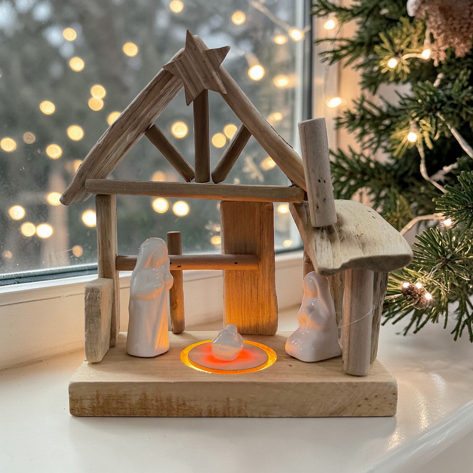 Holzkrippe mit Keramikfiguren auf einer Fensterbank platziert. Die integrierte LED-Beleuchtung leuchtet warm. Umgebung mit Tannenzweigen und Lichterkette, weihnachtliche Dekoration im Hintergrund.