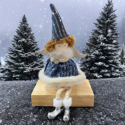 Sitzende Stoff-Engelfigur mit blauen Paillettenkleid, weißen Flügeln und Mütze auf einem Holzblock, im Hintergrund verschneite Tannenbäume.
