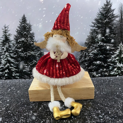 Sitzende Stoff-Engelfigur mit rotem Paillettenkleid, goldenen Flügeln und Mütze auf einem Holzblock, im Hintergrund verschneite Tannenbäume.