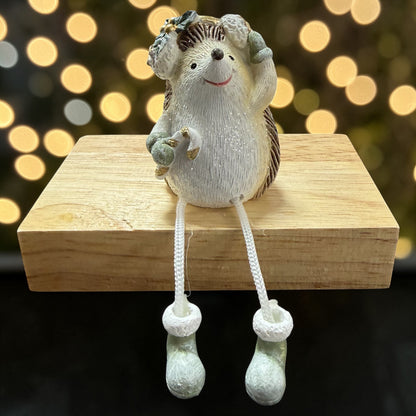 Eine Dekofigur in Form eines Igels in Weiß und Braun mit grünen Details und kleiner Zuckerstange, sitzend auf einem Holzblock vor dunklem Hintergrund mit warmen, runden Lichtreflexen.