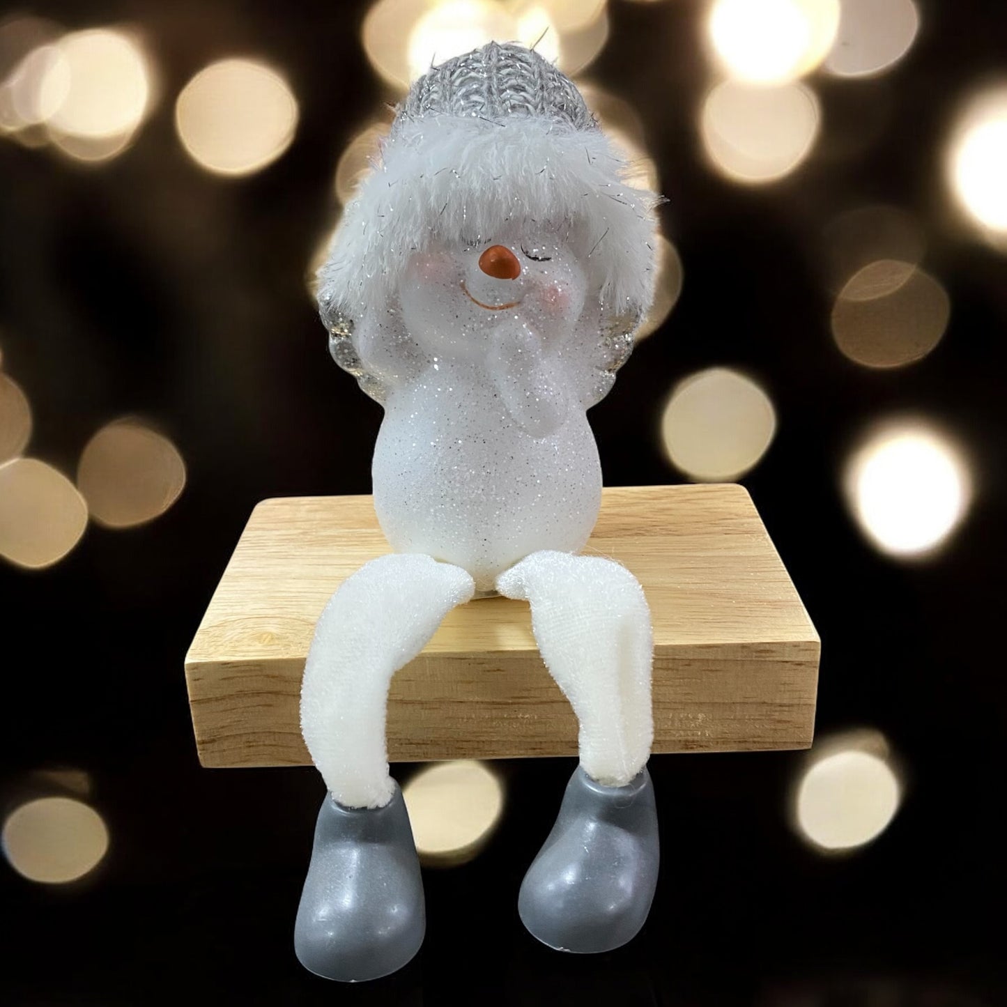 Sitzende Schneemannfigur mit grauer Mütze und weißen Beinen auf einem Holzblock, Hände am Kopf, vor unscharfem Hintergrund mit Lichtern.