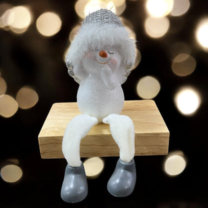 Sitzende Schneemannfigur mit grauer Mütze und weißen Beinen auf einem Holzblock, Hände am Kopf, vor unscharfem Hintergrund mit Lichtern.