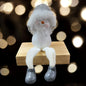 Sitzende Schneemannfigur mit grauer Mütze und weißen Beinen auf einem Holzblock, Hände am Kopf, vor unscharfem Hintergrund mit Lichtern.
